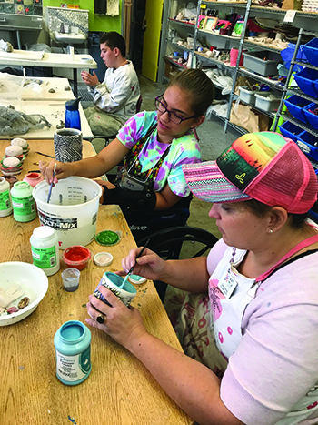 A woman and two students with disabilities paint while seated at a long table