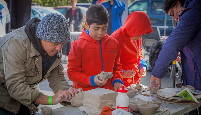 A man in wool cap and a young boy in a red jacket work with clay set out on a table