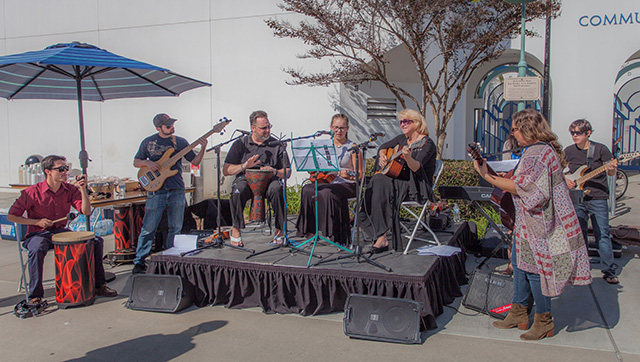 A man, older woman, and young girl perform on musical instruments on a small stage on a sunny day