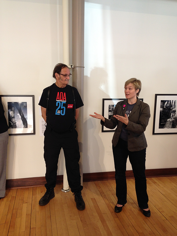 Man wearing ADA/25 shirt standing next to woman explaining exhibit. 