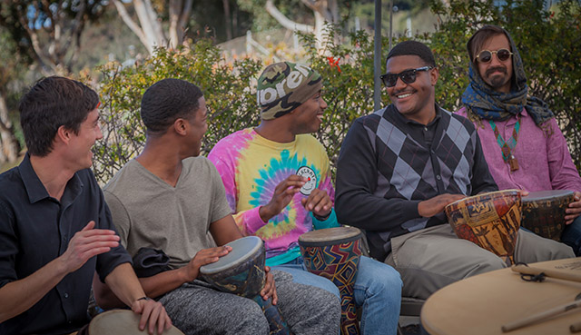 Young men sit side by side on chairs while playing hand drums outside