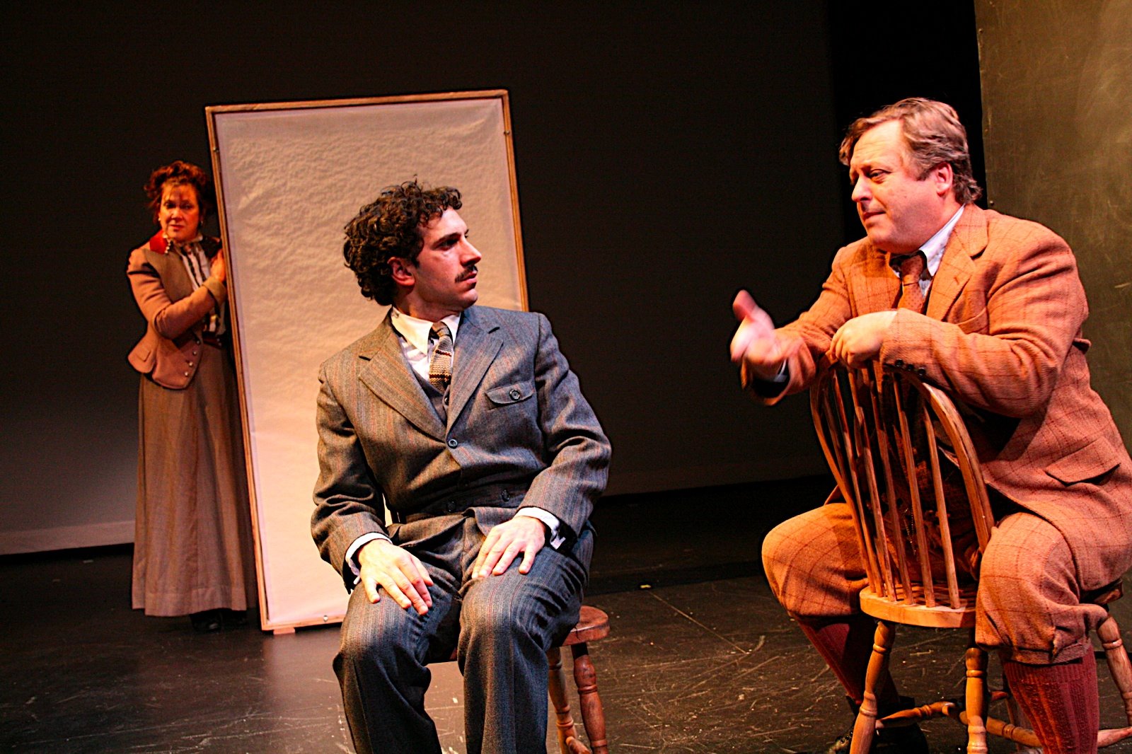 Two men sitting in wooden chairs talking, a woman listening behind a screen. 