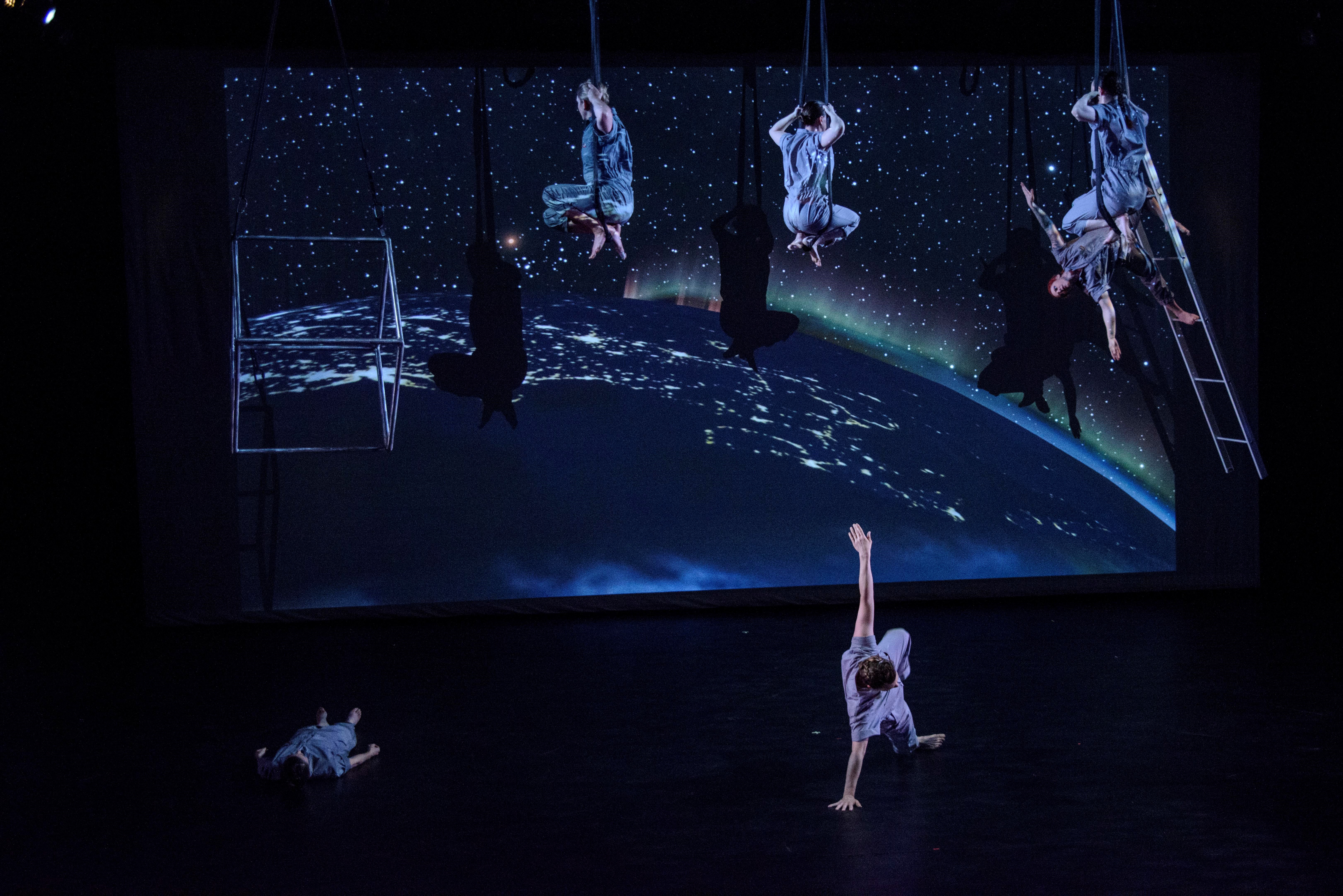 A group of performers on stage in dancing pose; some dancers are hanging in the air by an apparatus. 