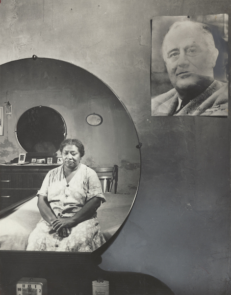 portrait of woman reflected in a circular mirror as she sits on her bed with framed portrait of Franklin D. Roosevelt in foreground of photo
