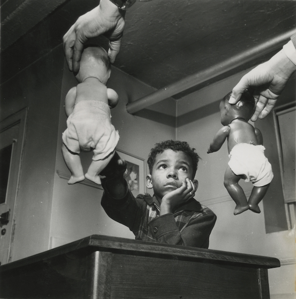 a young black boy sits at a table while the arms of someone out of the frame dangle two dolls in front of him