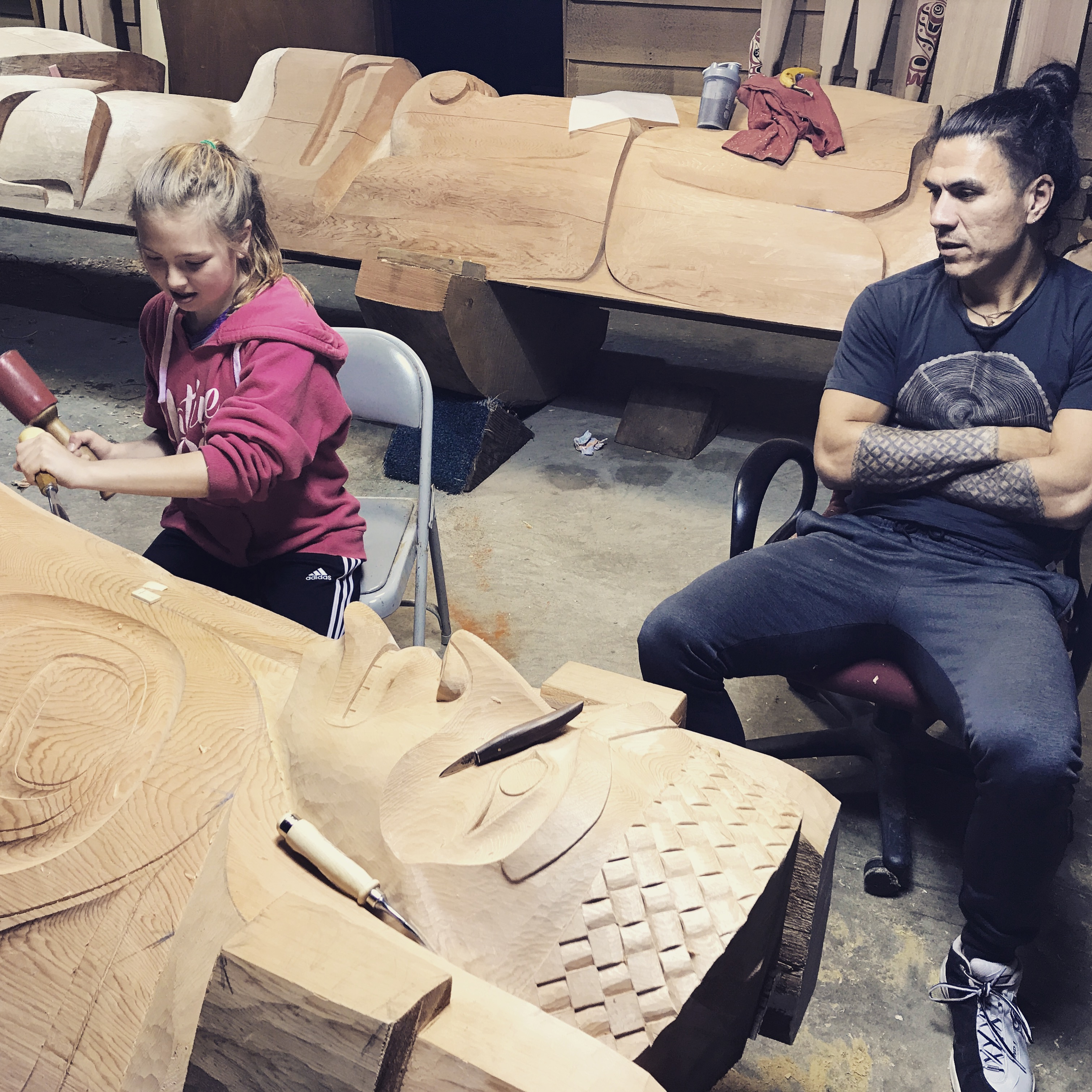 A young girl carves a totem pole while her father watches