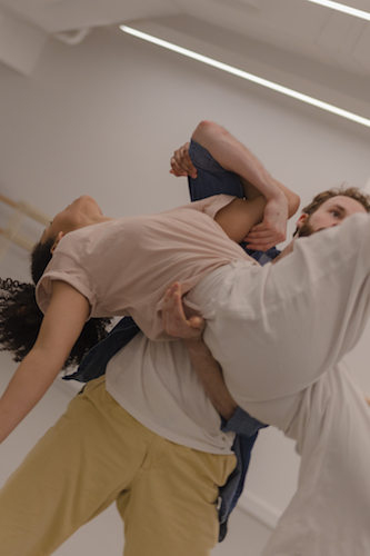 In a rehearsal studio, a male dancer holds a womn dancer suspended in air pressed close to his body
