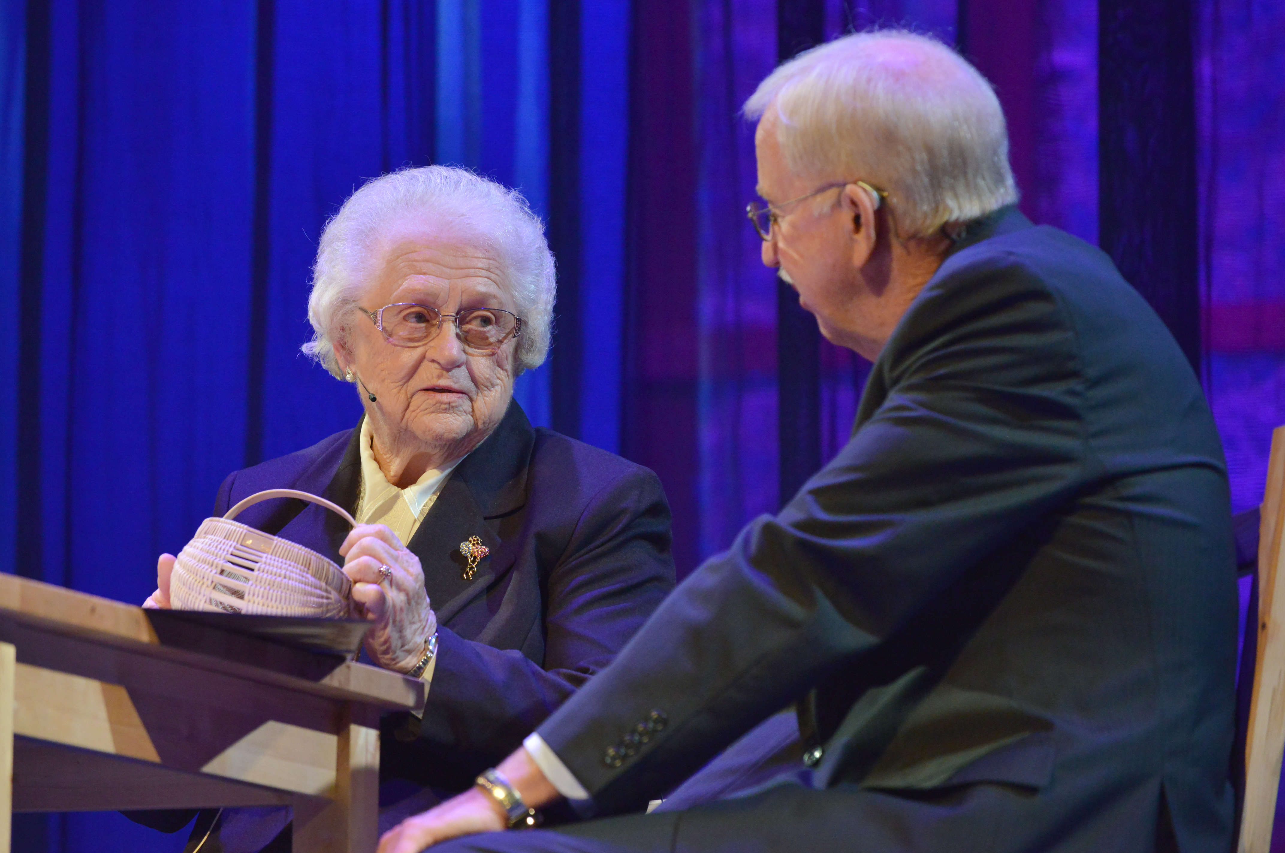 The fellow is sitting down with a basket in her hand and she is talking to the emcee.