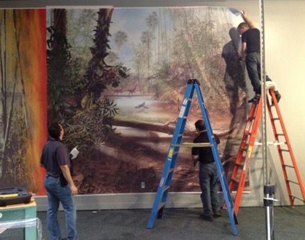 A man stands on a ladder as he installs a large mural of dinosaurs while two other men look on