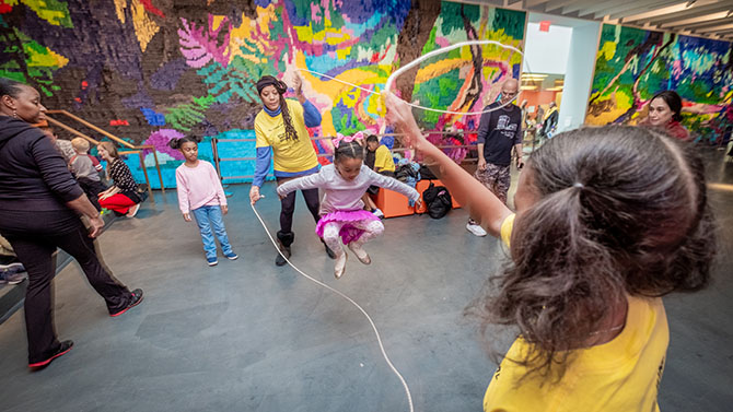 Children do double dutch jump rope inside in front of a large mural made of paper fringe