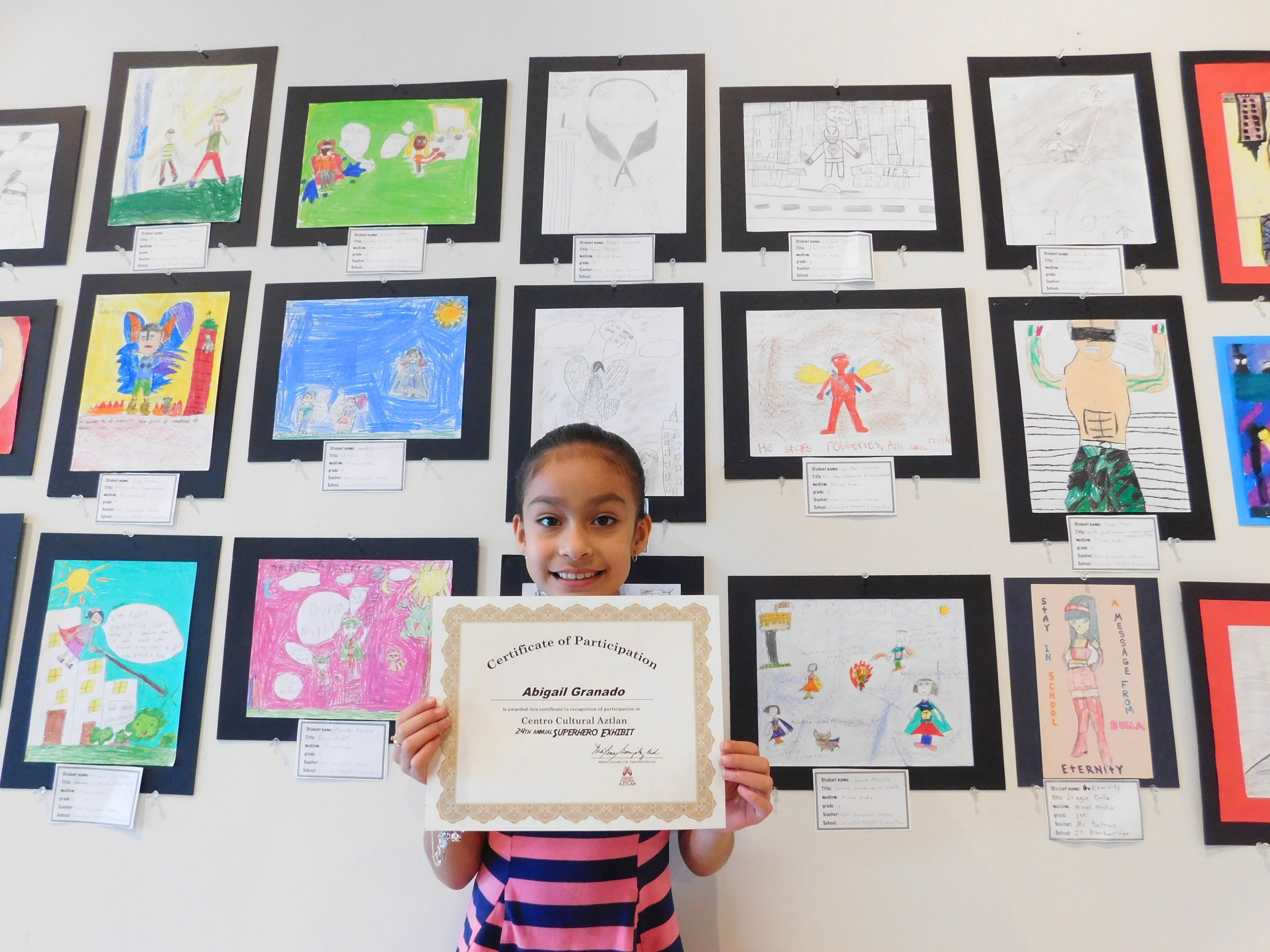 Young girl holding up a certificate of participation in front of a wall of drawings.
