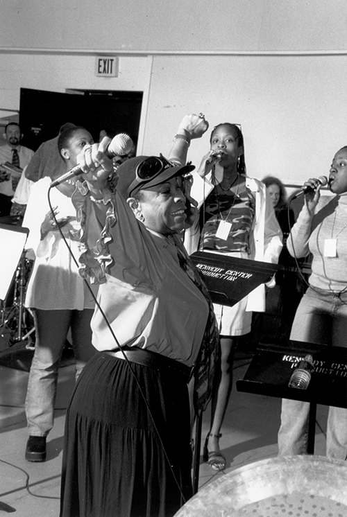 Woman wearing baseball cap and holding a microphone gesturing in front of a group of students. 