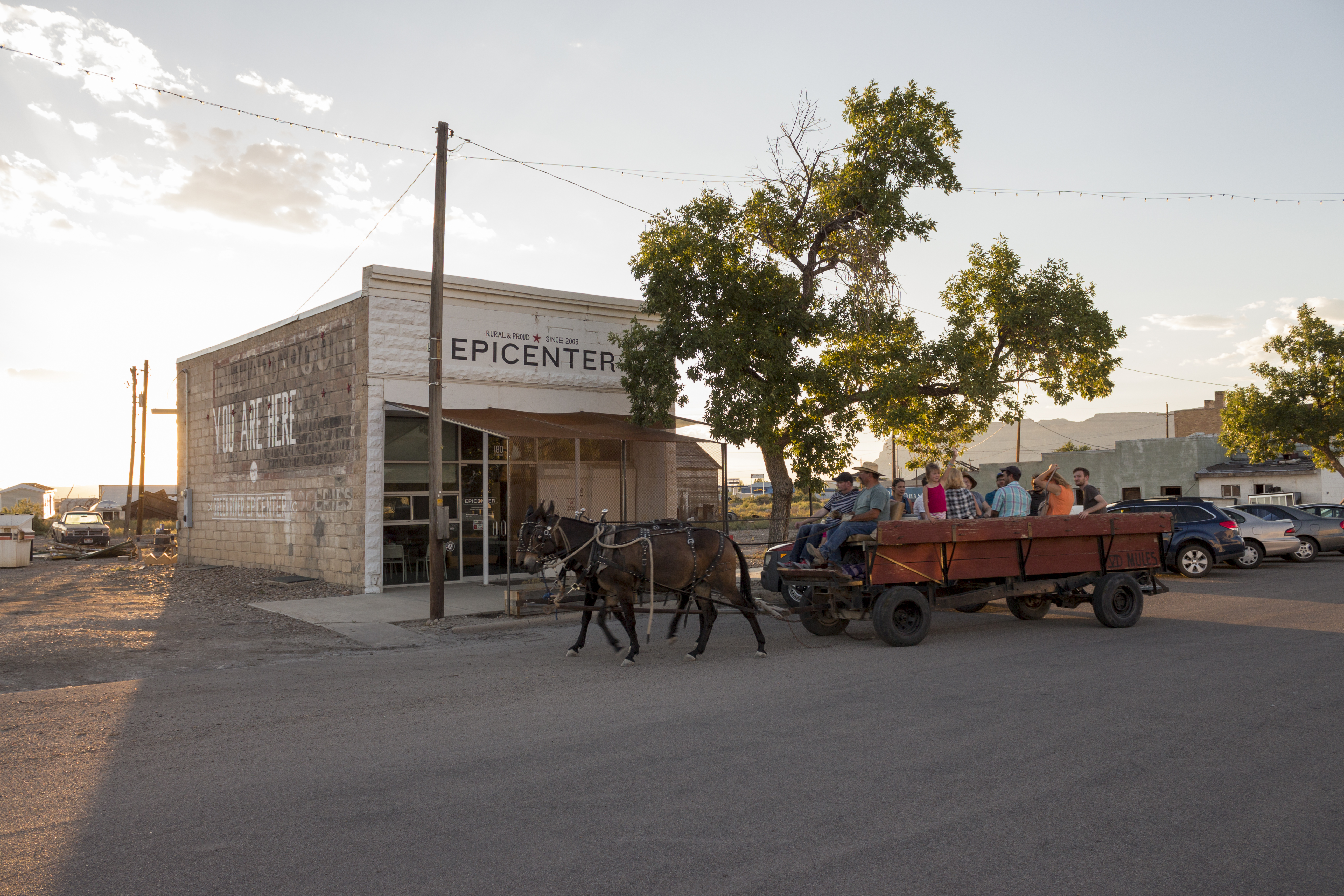 Group of people in a cart being pulled by horses in front of a small building. 