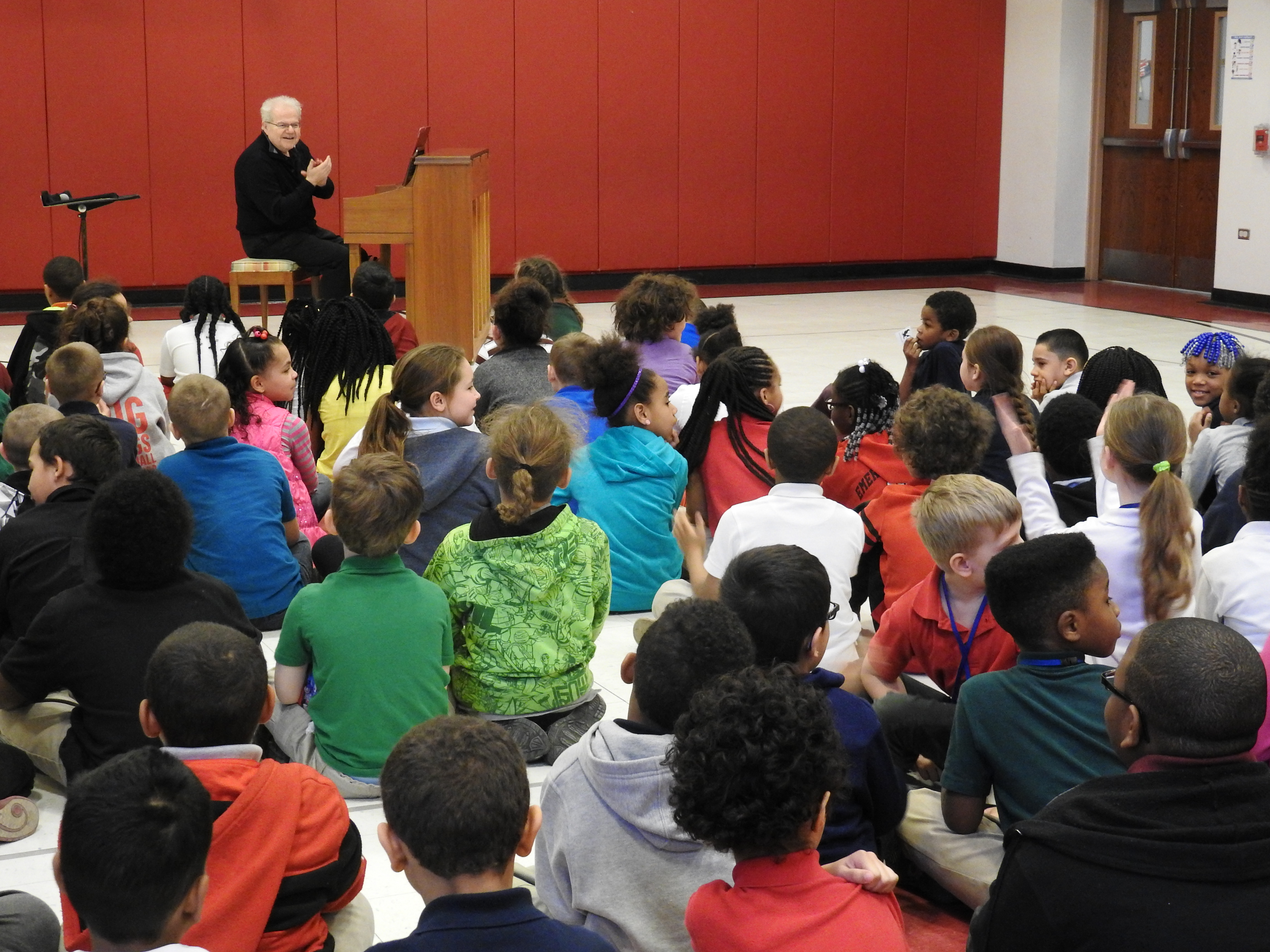 Man at piano speaking to a group of children sitting on the floor. 