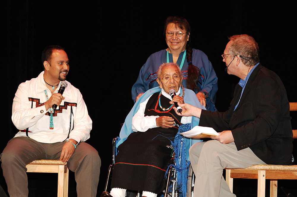 Elderly woman being interviewed onstage surrounded by her family. 