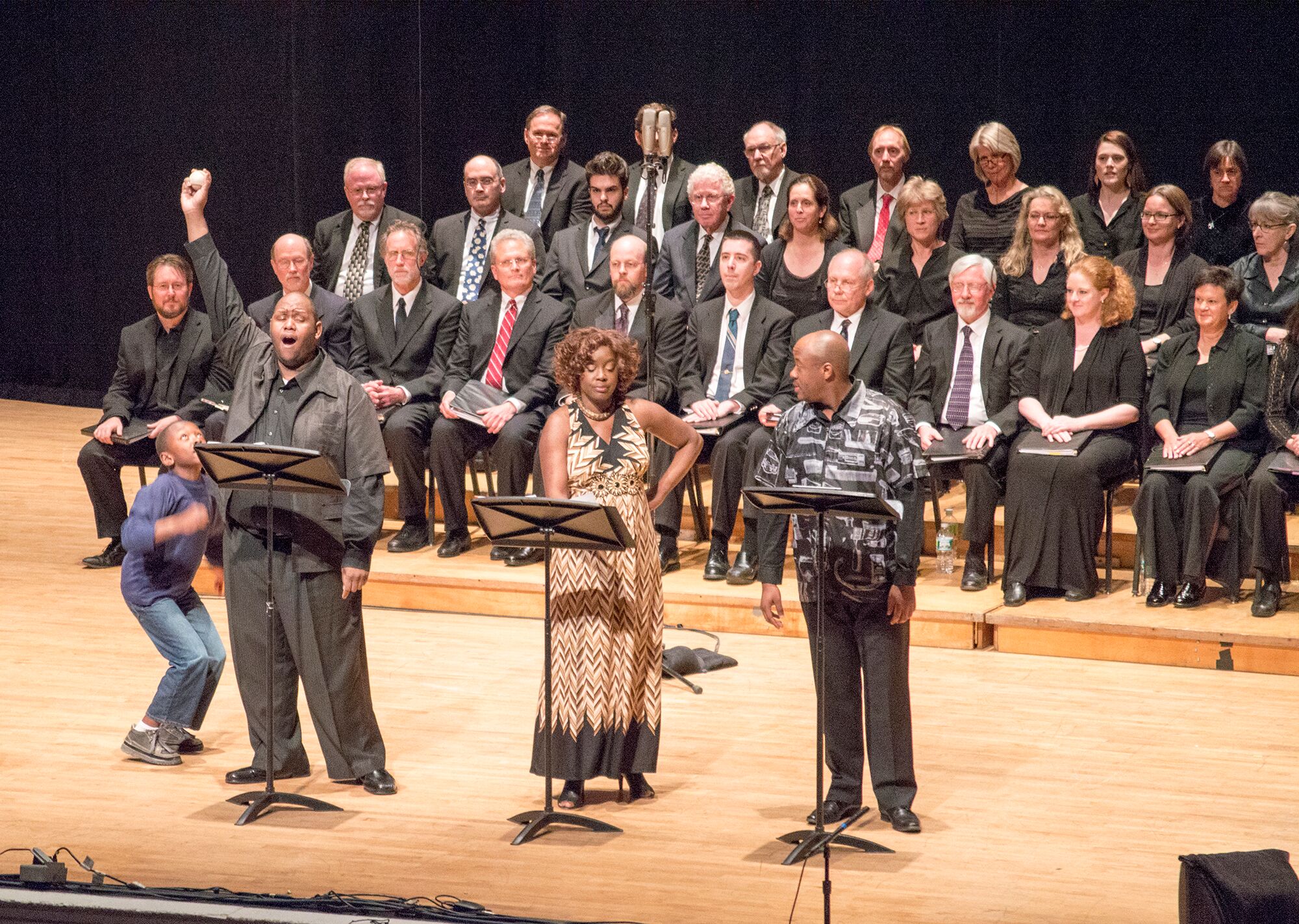 Three singers on stage, one holding a baseball, with a choir sitting behind them. 