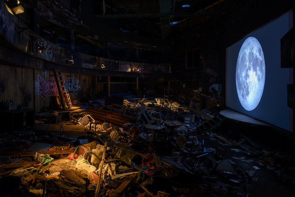 A room filled with junk in front of a screen showing the moon. 