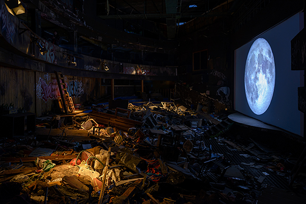 A room filled with junk in front of a screen showing the moon.