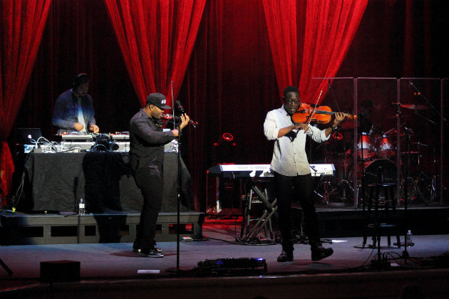 Two men play violin onstage in front of a DJ