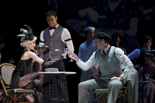 A woman and man in 1920s clothes sitting at a table with drinks in hand, waiter standing next to their table. 