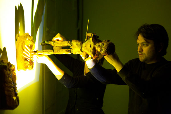 A man holds a wooden puppet up to a lit screen