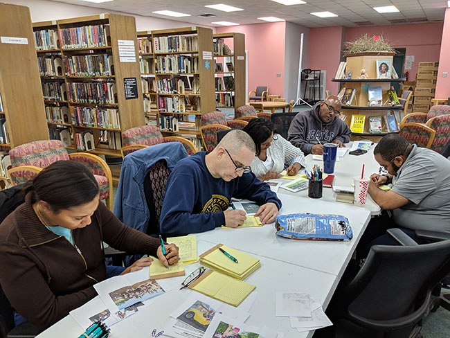 With library stacks in the background, men and women sit around a table and write on yellow legal pads