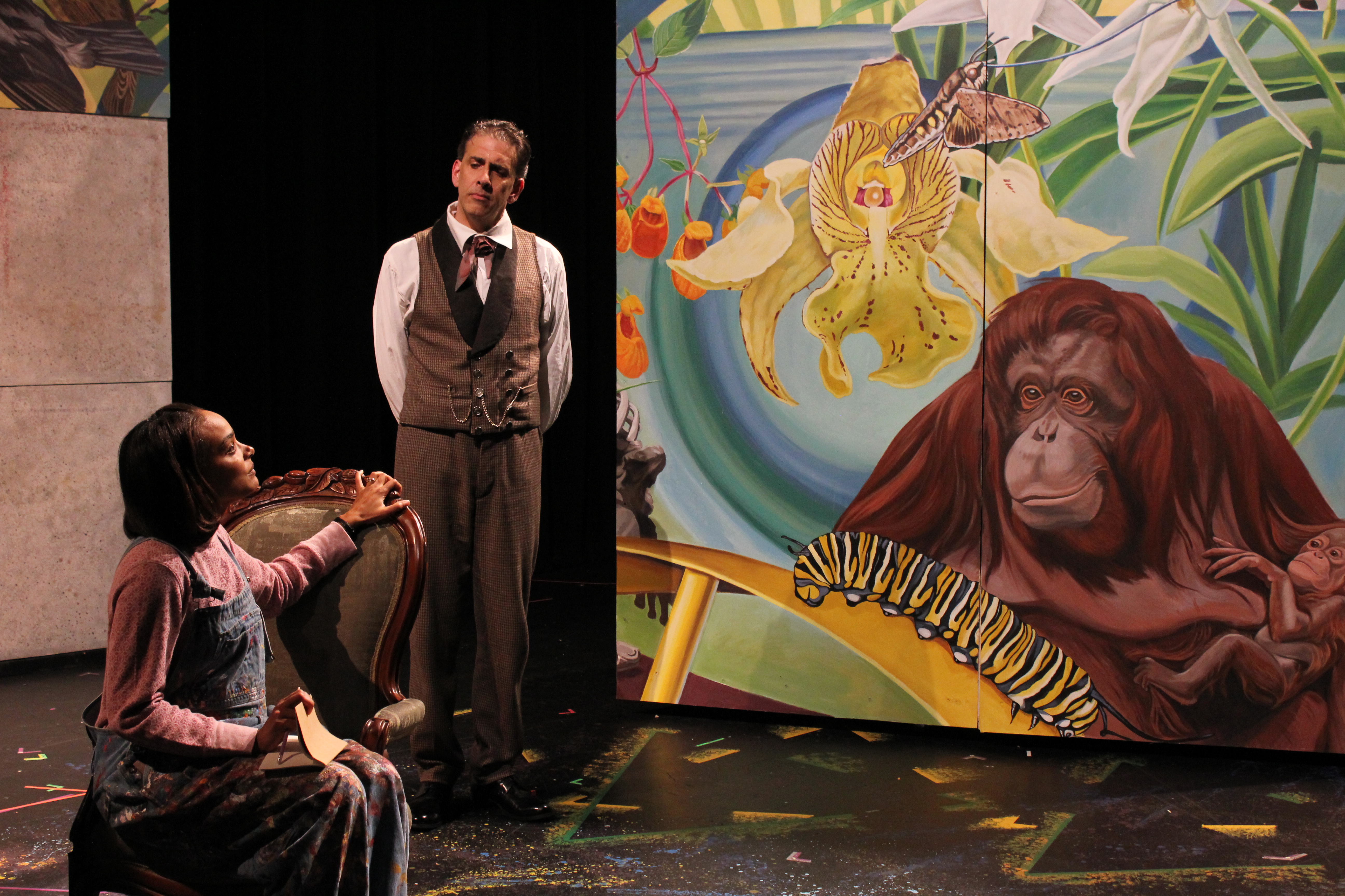 An African-American woman sitting in front of a white male, both in front of a mural showing a gorillia, catepillar, and butterfly. 