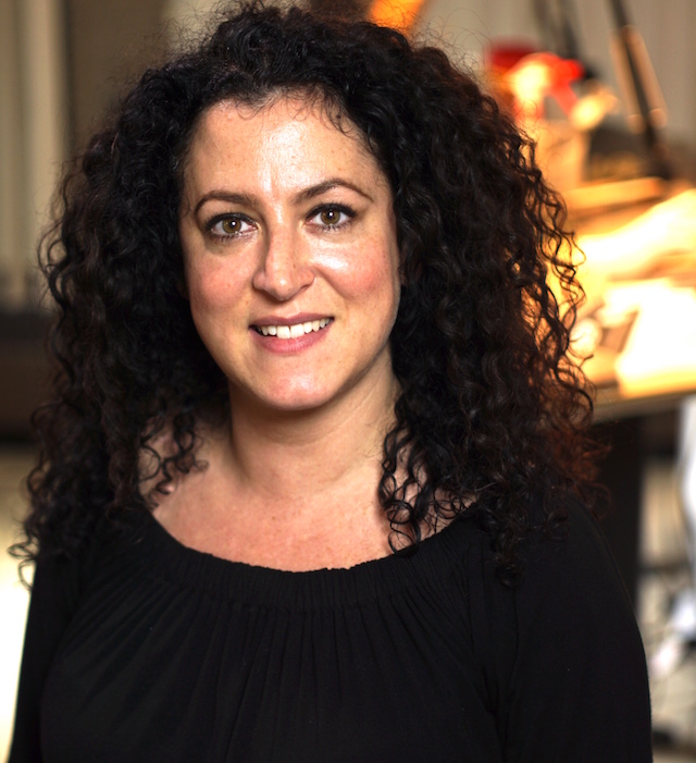 Renata Gordon who is a white woman with long dark curly hair. This is a close-up shot and you can see dim shapes of a workshop behind her her.