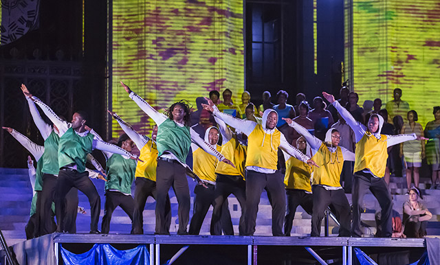 Teen boys in green and yellow shirts hold out their arms diagonally as they perform a step routine on the steps of the public library at night