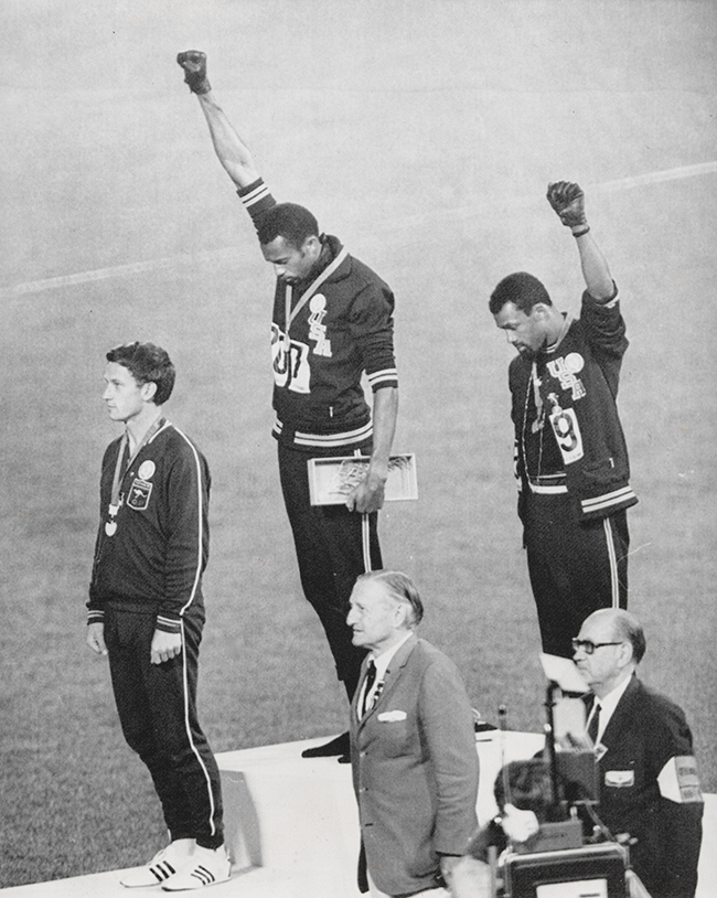 Two men raise their fists while standing on platforms during the Olympics medals ceremony