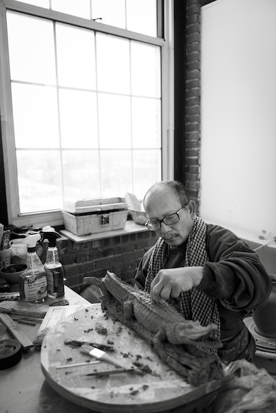 An older Cambodian man in glasses is carving a small ceramic alligator figurine