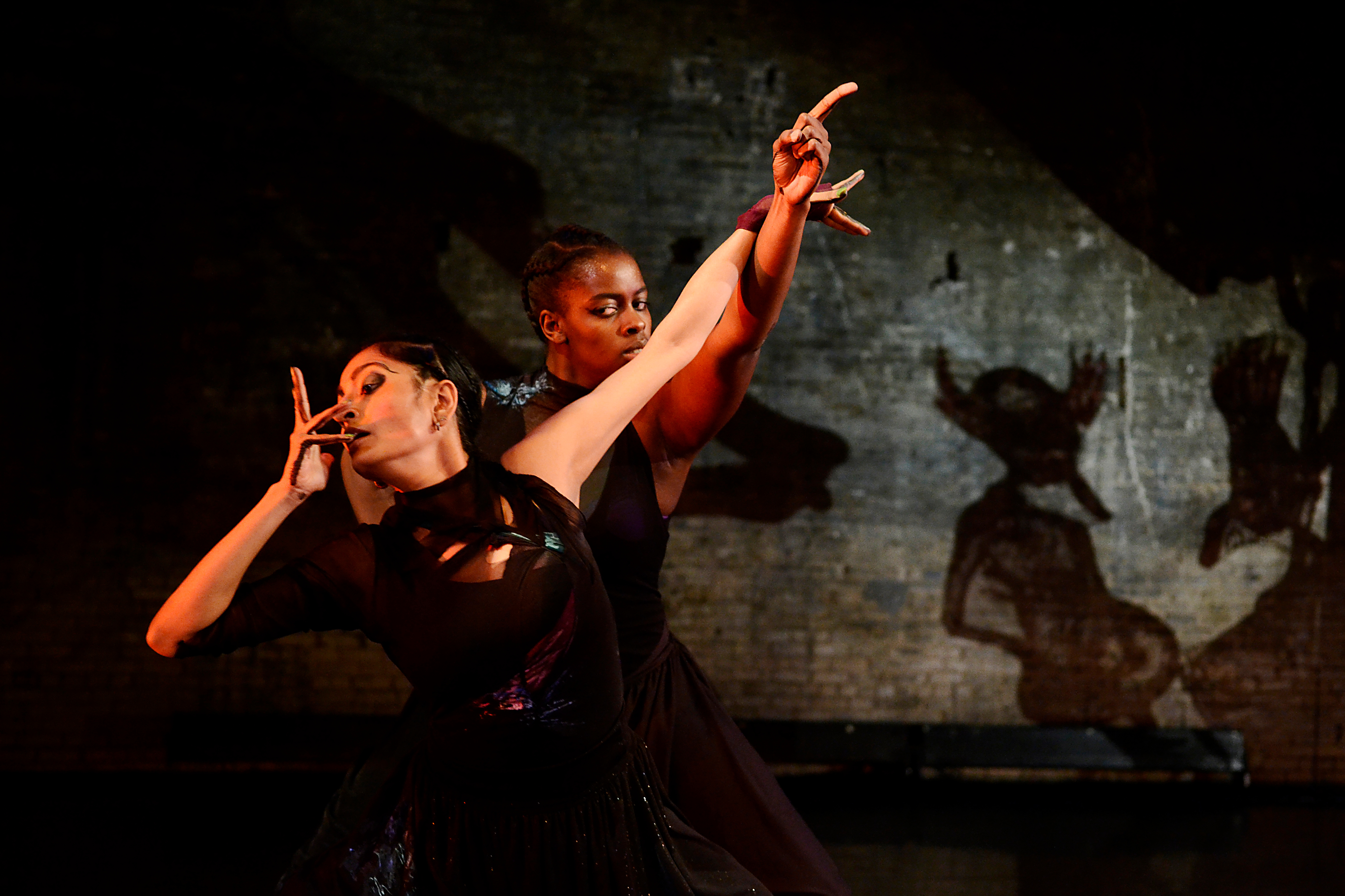 Tdwo female dancers posing together; one standing in front of the other, both in dark attire and on a dark stage setting.