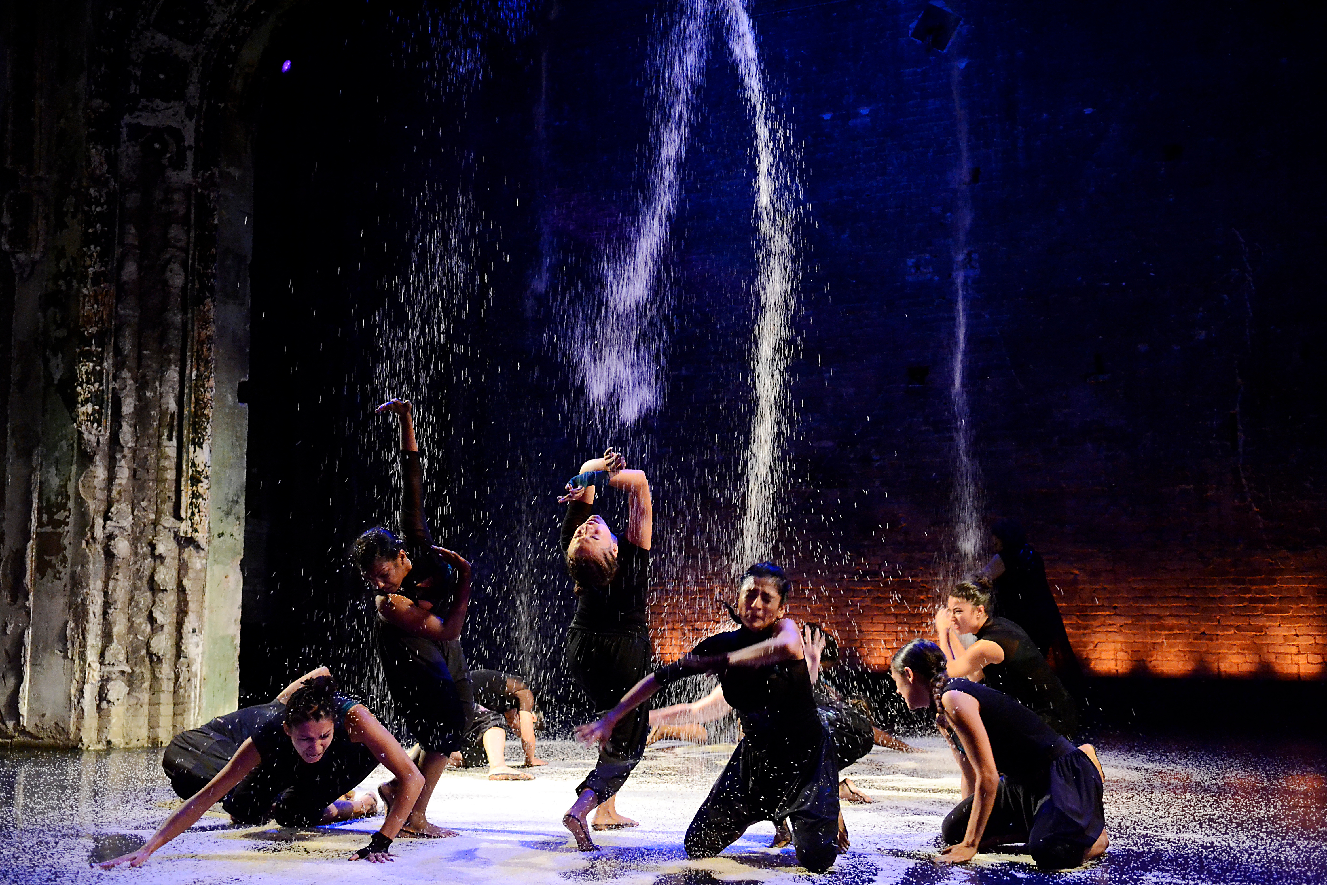 A group of female dancers on stage pounding the stage as water falls on them.