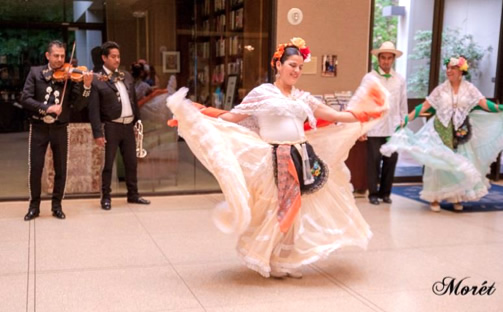 two female dancers in traditional Mexican costume perform. Two mariachi musicians play behind.