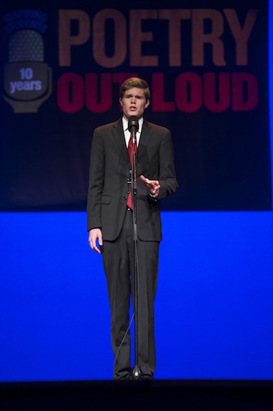 a very tall slender young white man in a suit recites a poem into a microphone