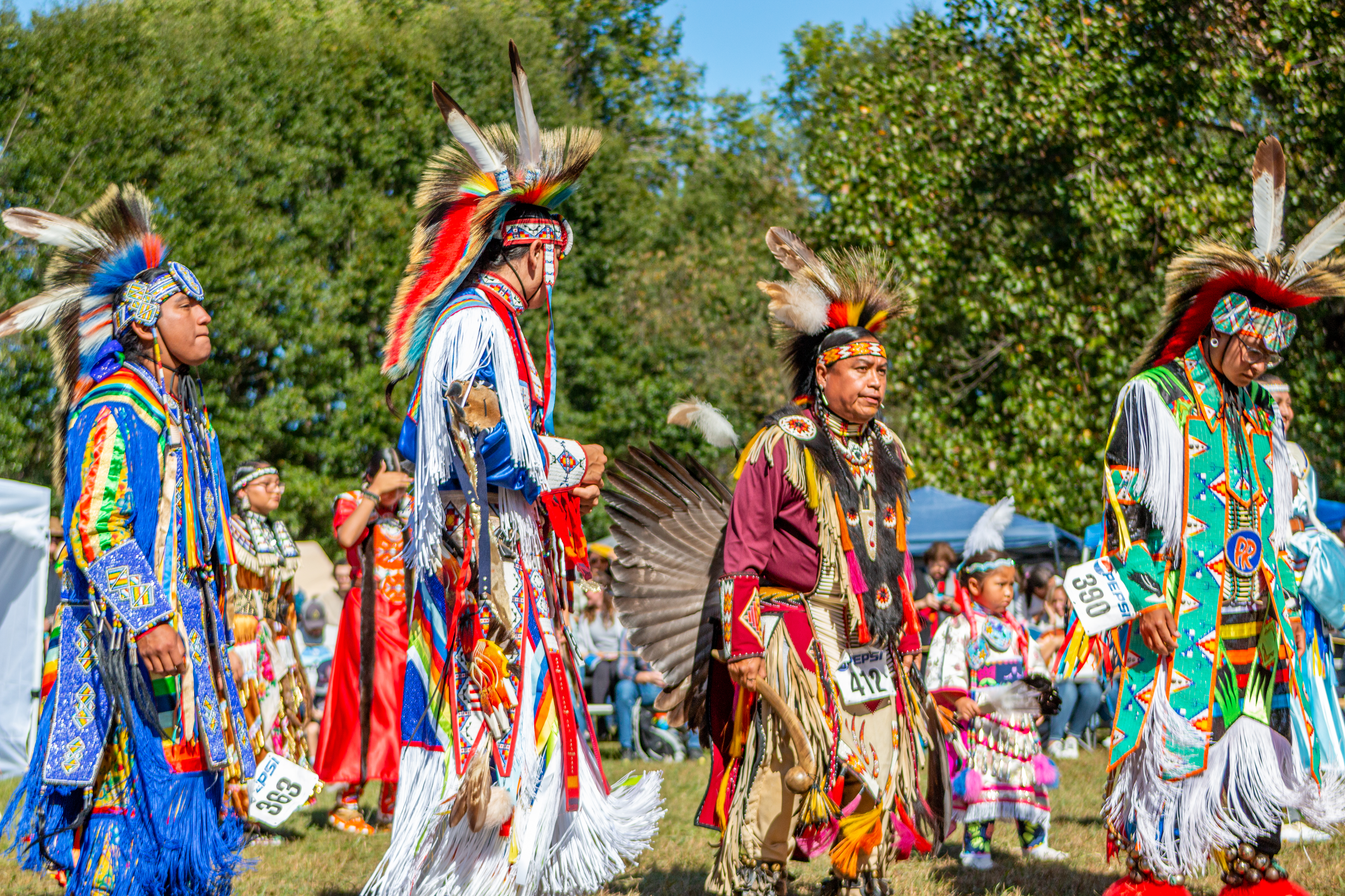 A group of four men in traditional regalia walking in a line outdoors.