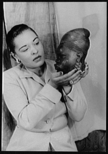 black and white photo of Billie Holiday who is shown from the waist up and stands holding an African mask