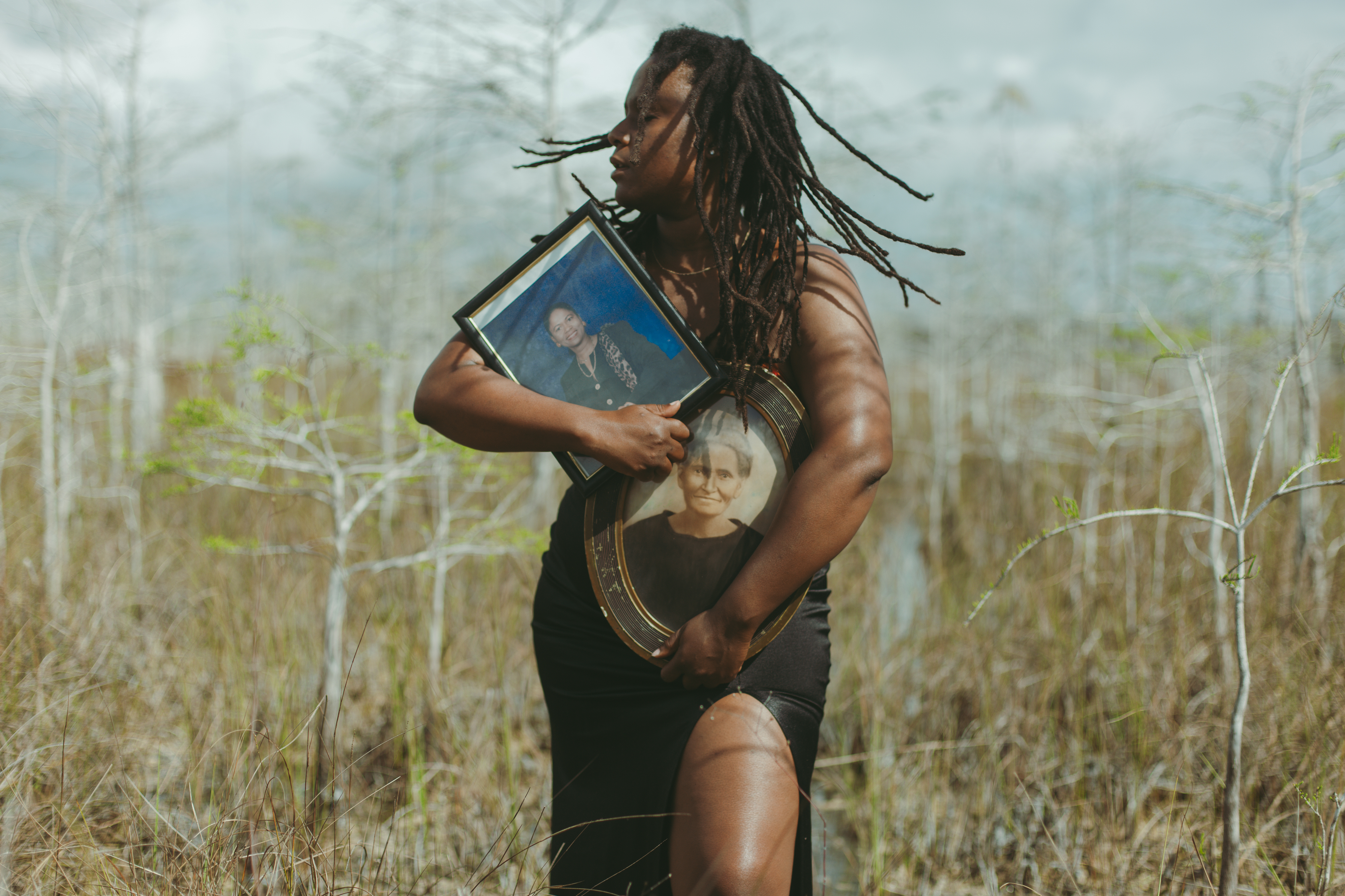 A Black woman holds two photos in vintage frames in the midst of a swamp landscape