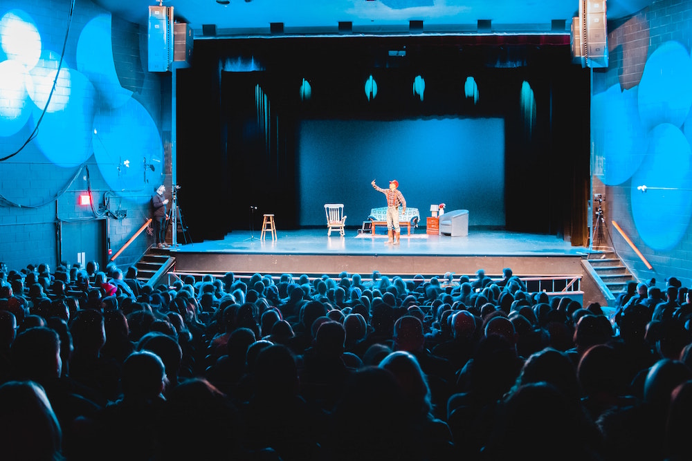 an actor performs on a blue-lit stage; you can see the audience watching him