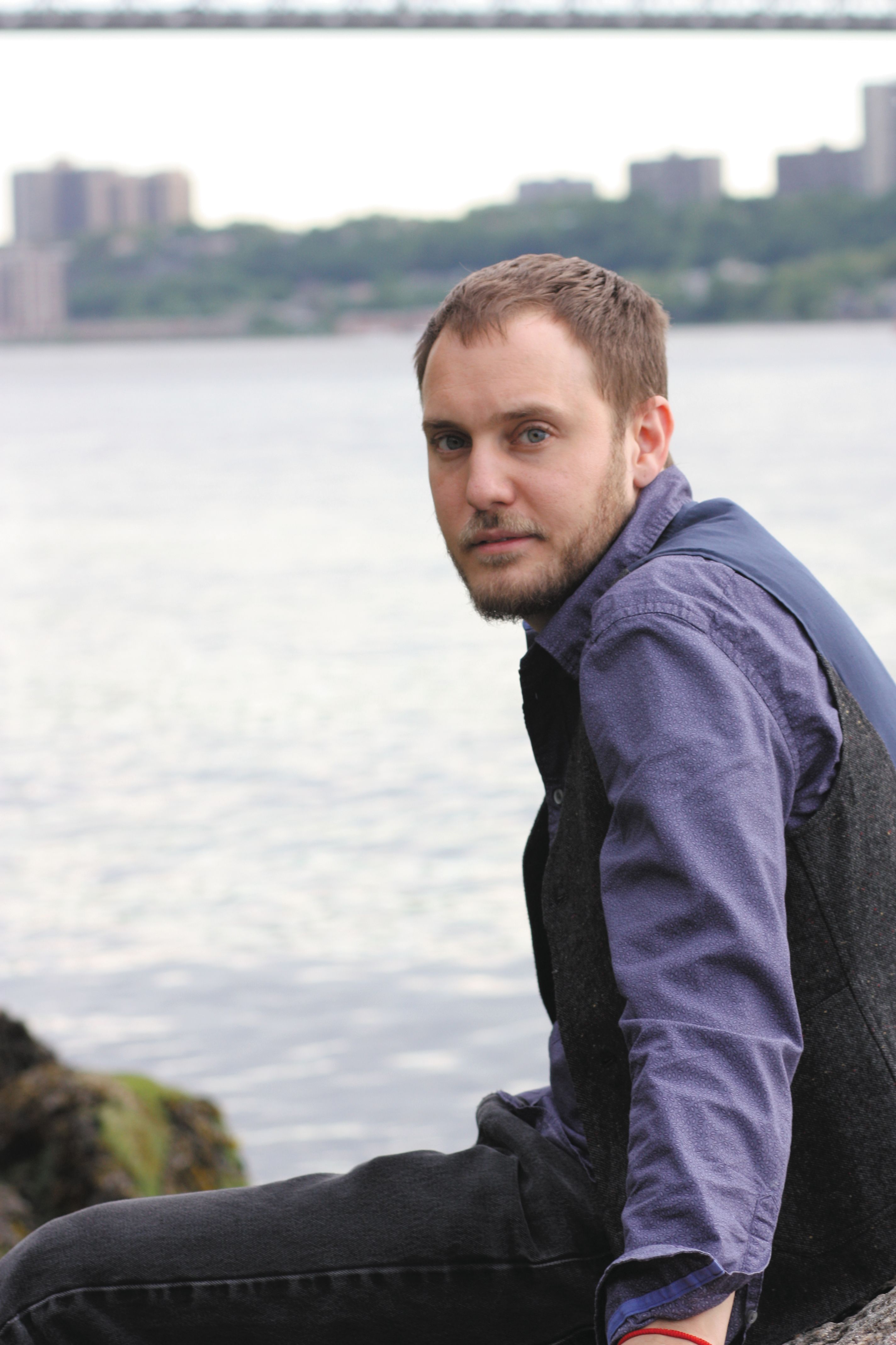 A white man with navy blue shirt and a black vest poses at a waterfront.