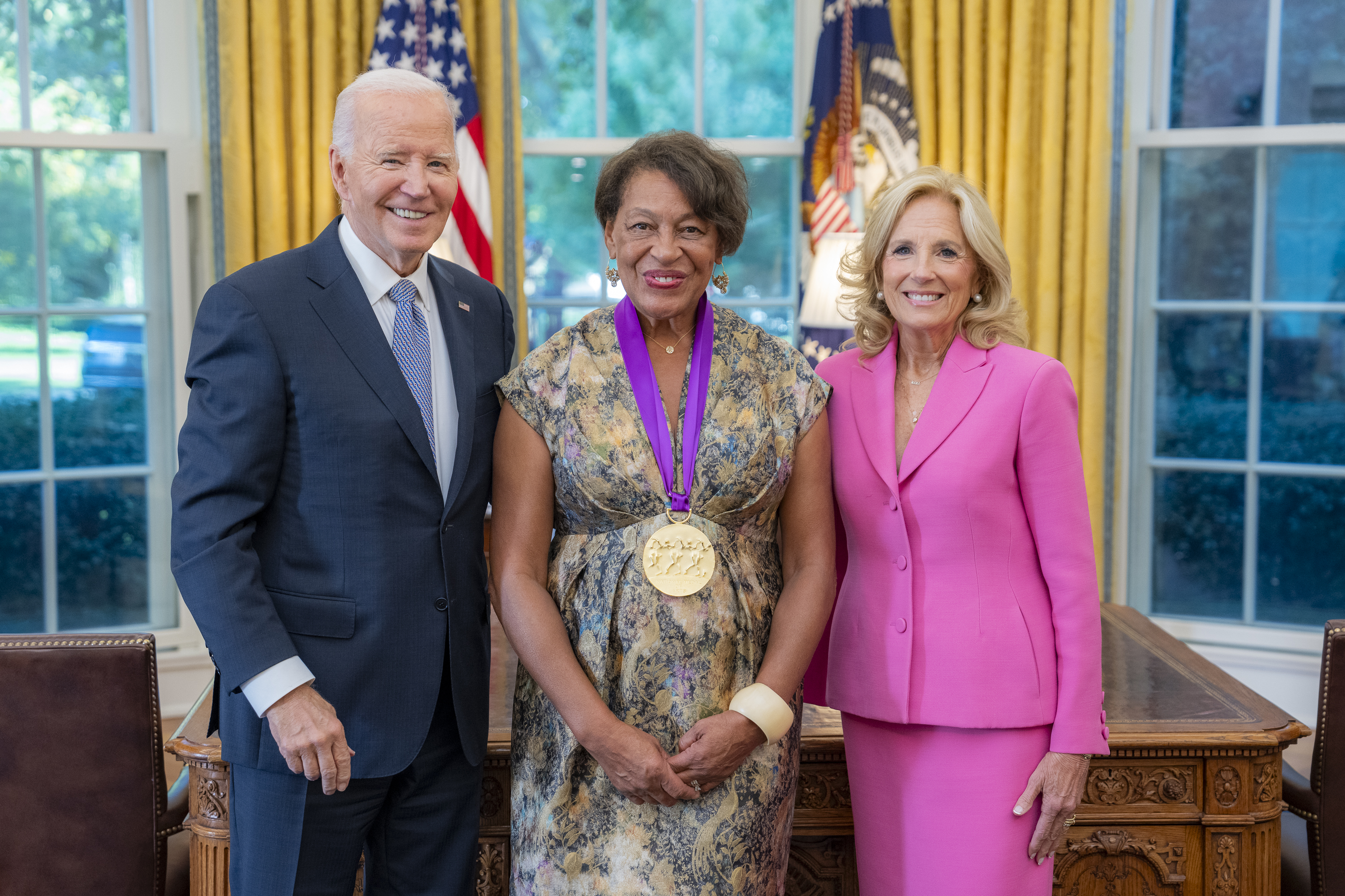 Black woman in flowery dress wearing a medal around her neck surrounded by older man wearing a blue suit and woman with blonde hair wearing a pink suit.
