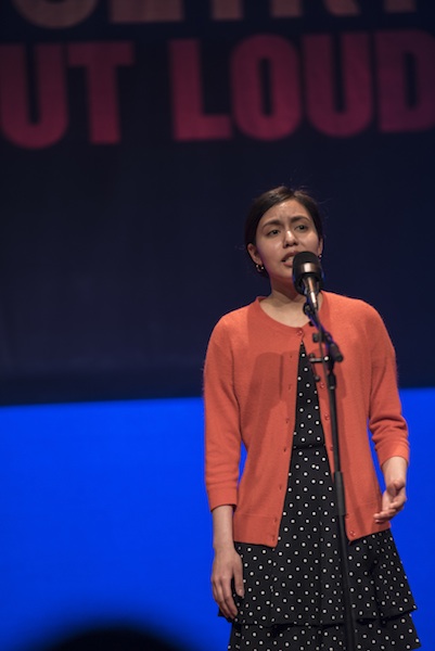 A young woman stands on stage reciting into a microphone