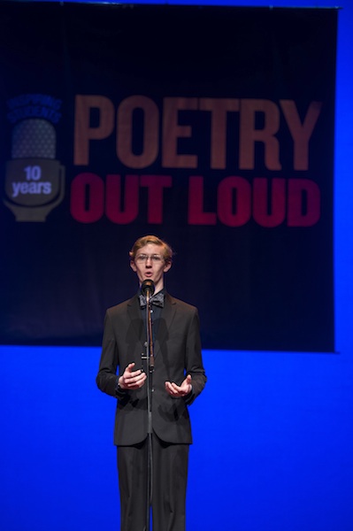 a tall young man in a suit and bowtie recites on stage into a microphone