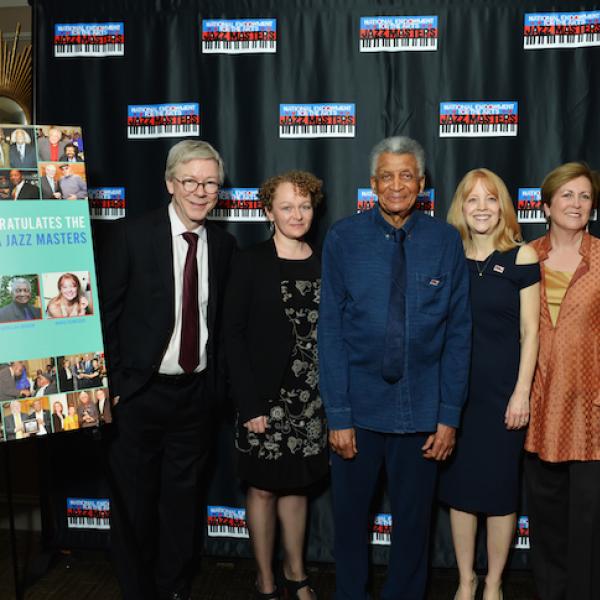 Six people stand smiling in front of a backdrop with the NEA Jazz Masters logo repeated. Next to them on an easel is a poster saying “BMI Congratulates the 2019 NEA Jazz Masters” with photos.