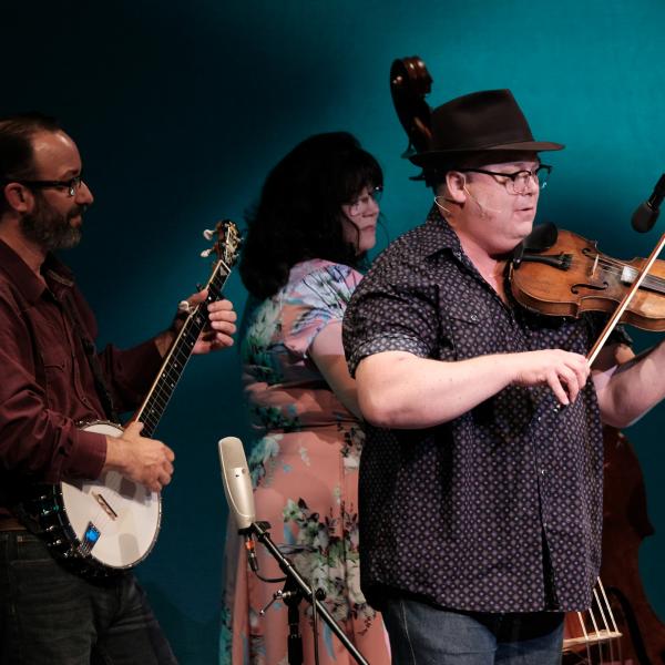 A man stands in front of a microphone playing the fiddle while behind him are musicians playing the banjo, bass, and guitar.