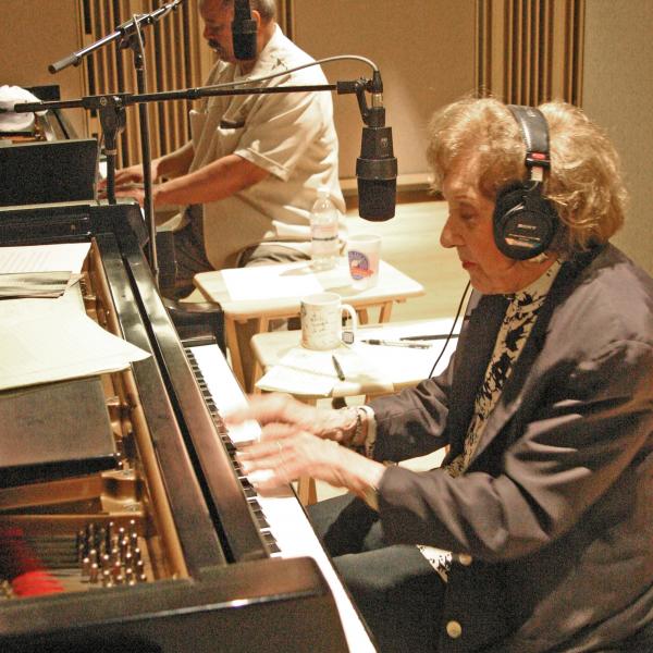 Woman wearing headphones playing piano with African-American man behind her playing piano.