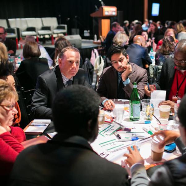 A group of men and women sit around a round table listening to one person at the table speak.