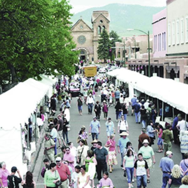 Crowds of people walking down the street and looking into various white tents lined up along the curb; an old church in the background.