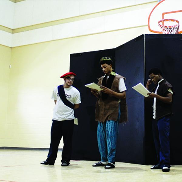 Three young men wearing costumes read from scripts in a gymnasium