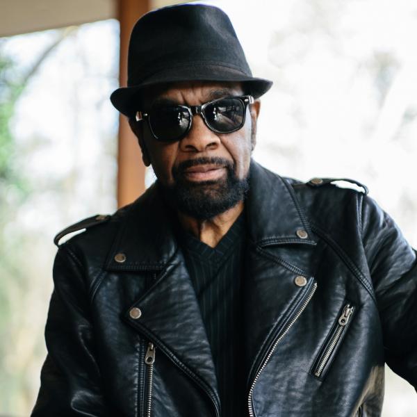 Man wearing a pork pie hat, sunglasses, and a leather jacket. 